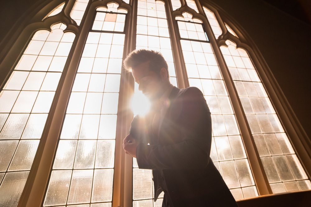 Groom silhouetted in sunlight at a tall church window while fastening his jacket