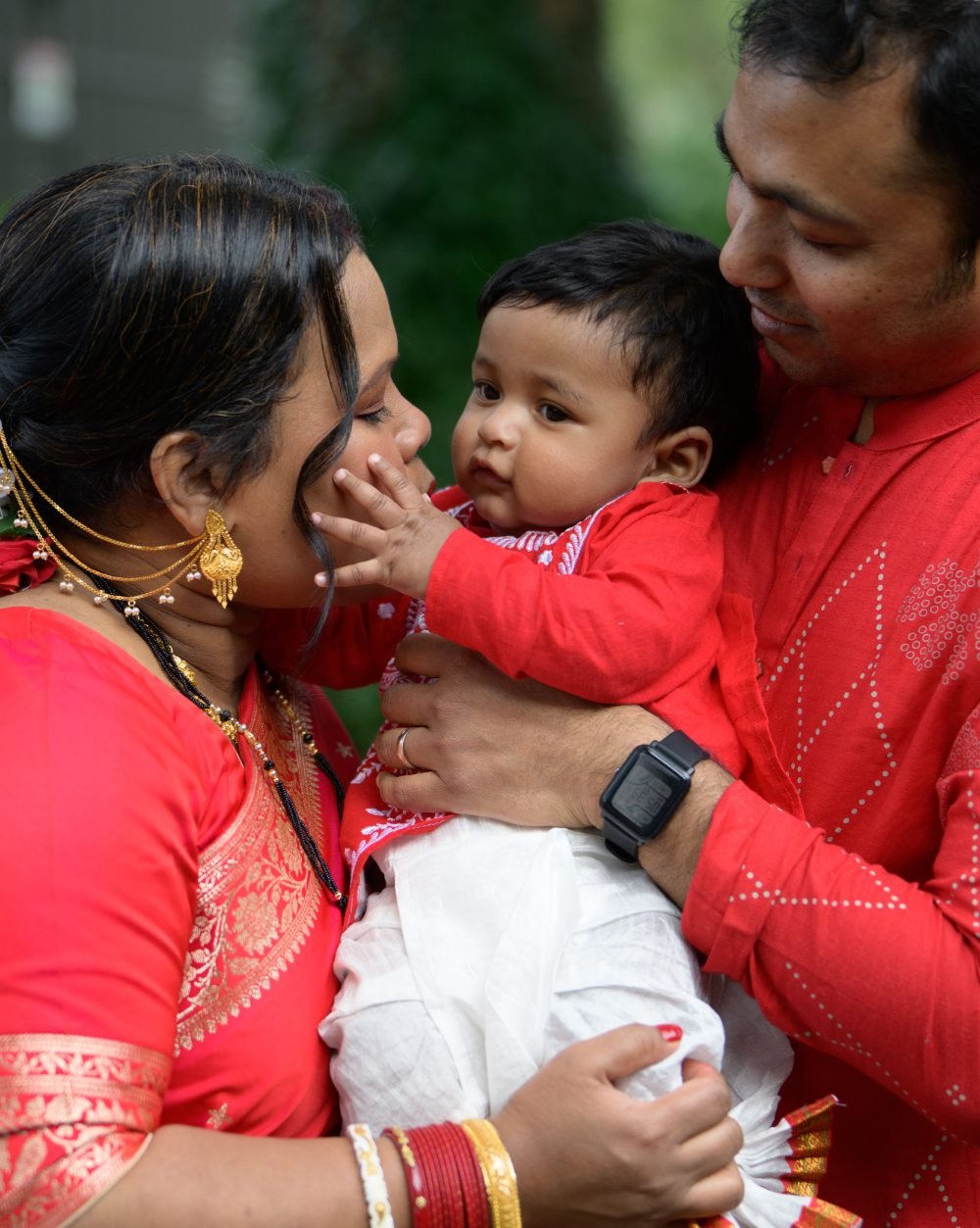 Parents in red traditional dress holding their baby during a family celebration outdoors