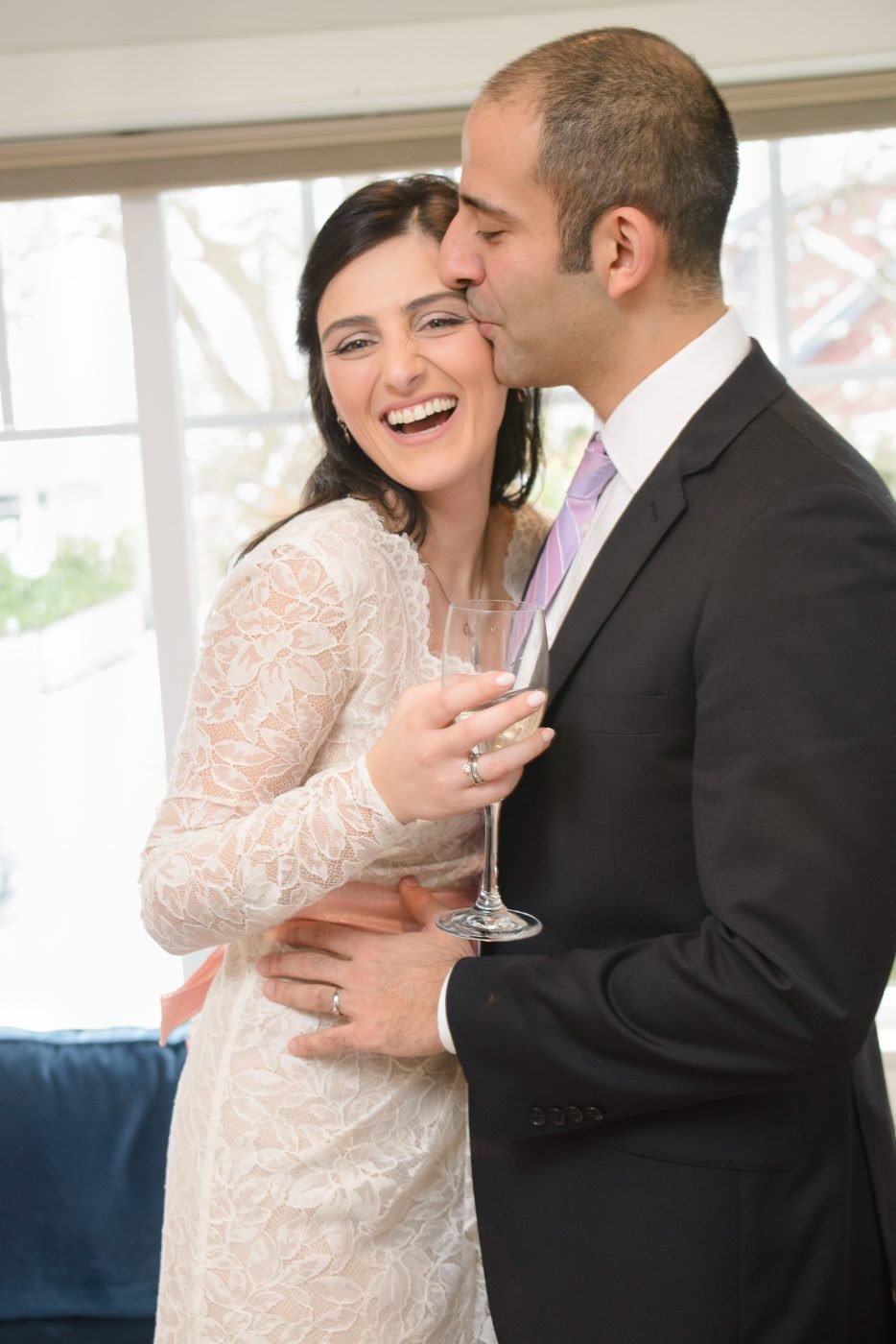 Groom kissing his laughing bride on the cheek during a champagne toast at their wedding reception, photographed in the Lower Mainland