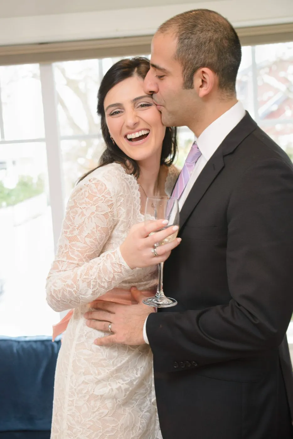 Groom kissing his laughing bride on the cheek during a champagne toast at their wedding reception, photographed in the Lower Mainland