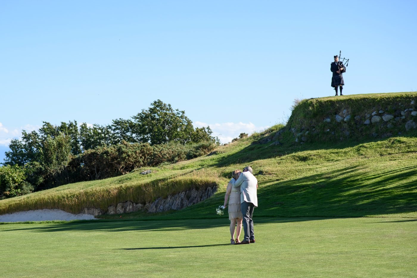 Wedding couple embracing on a golf course fairway while a bagpiper plays on the hill above them