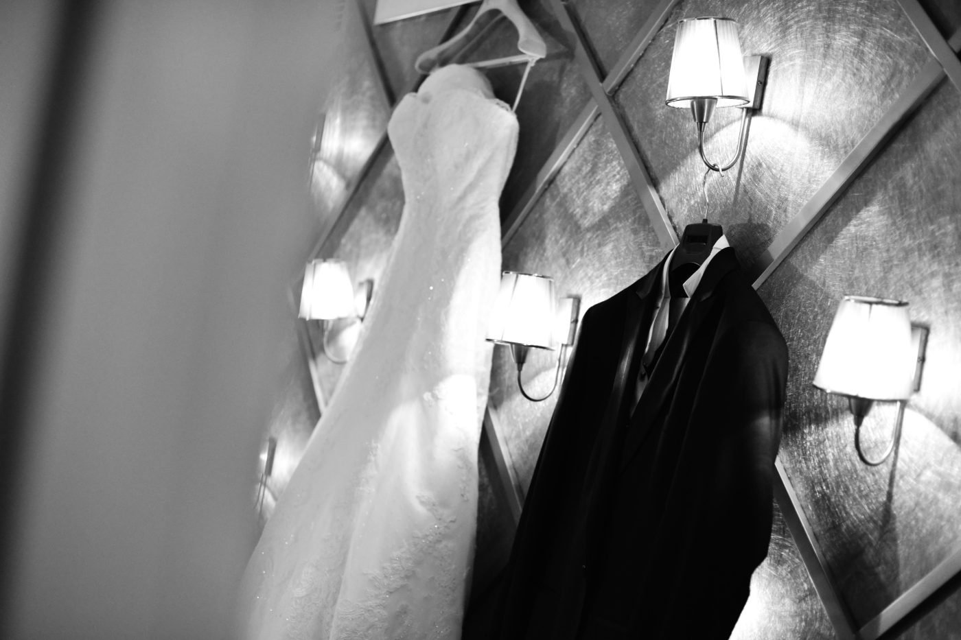 Black and white getting ready detail of the wedding dress and the groom&rsquo;s suit hanging side by side at the venue
