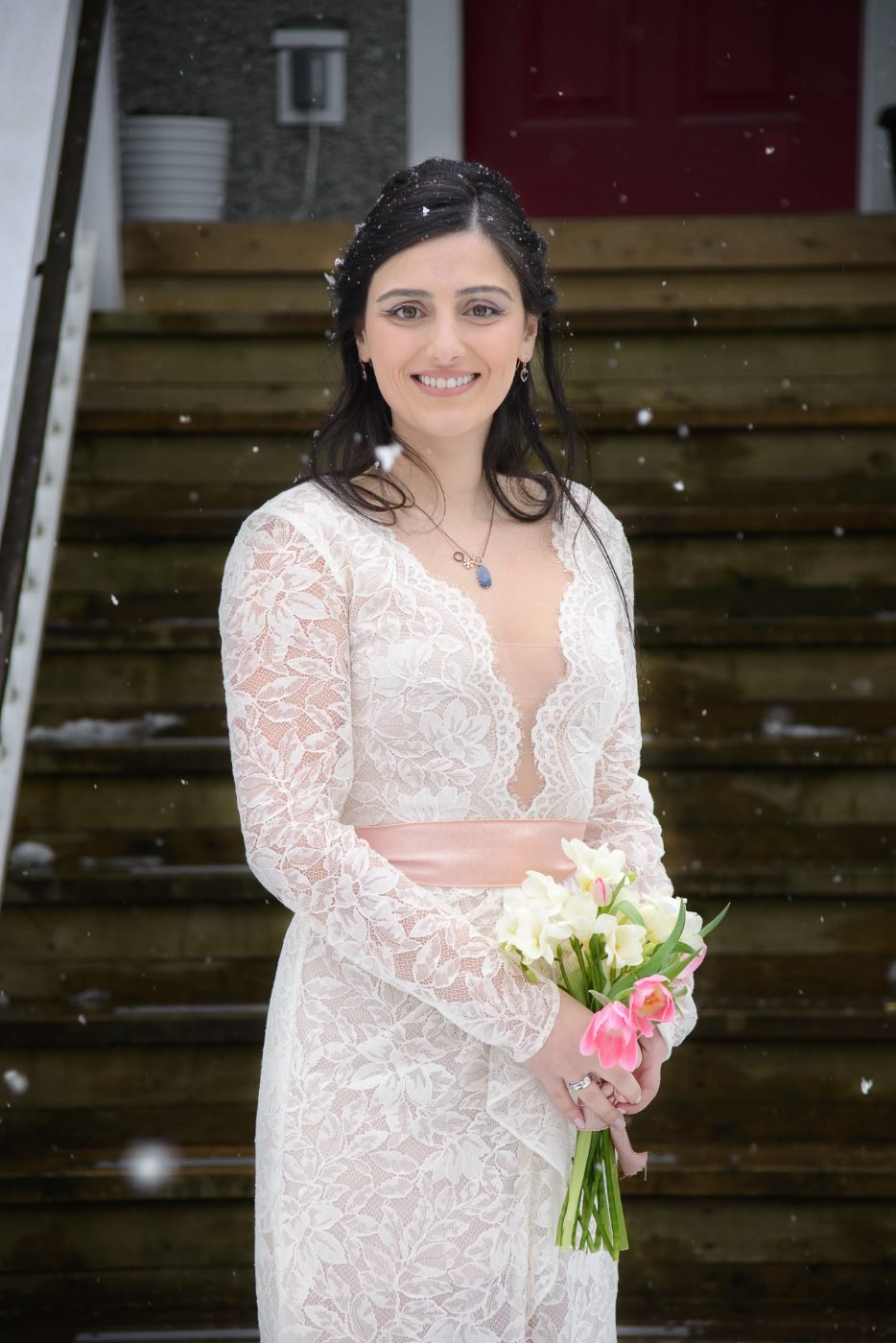 Winter bride portrait in a lace gown holding tulips as snow falls on the front steps