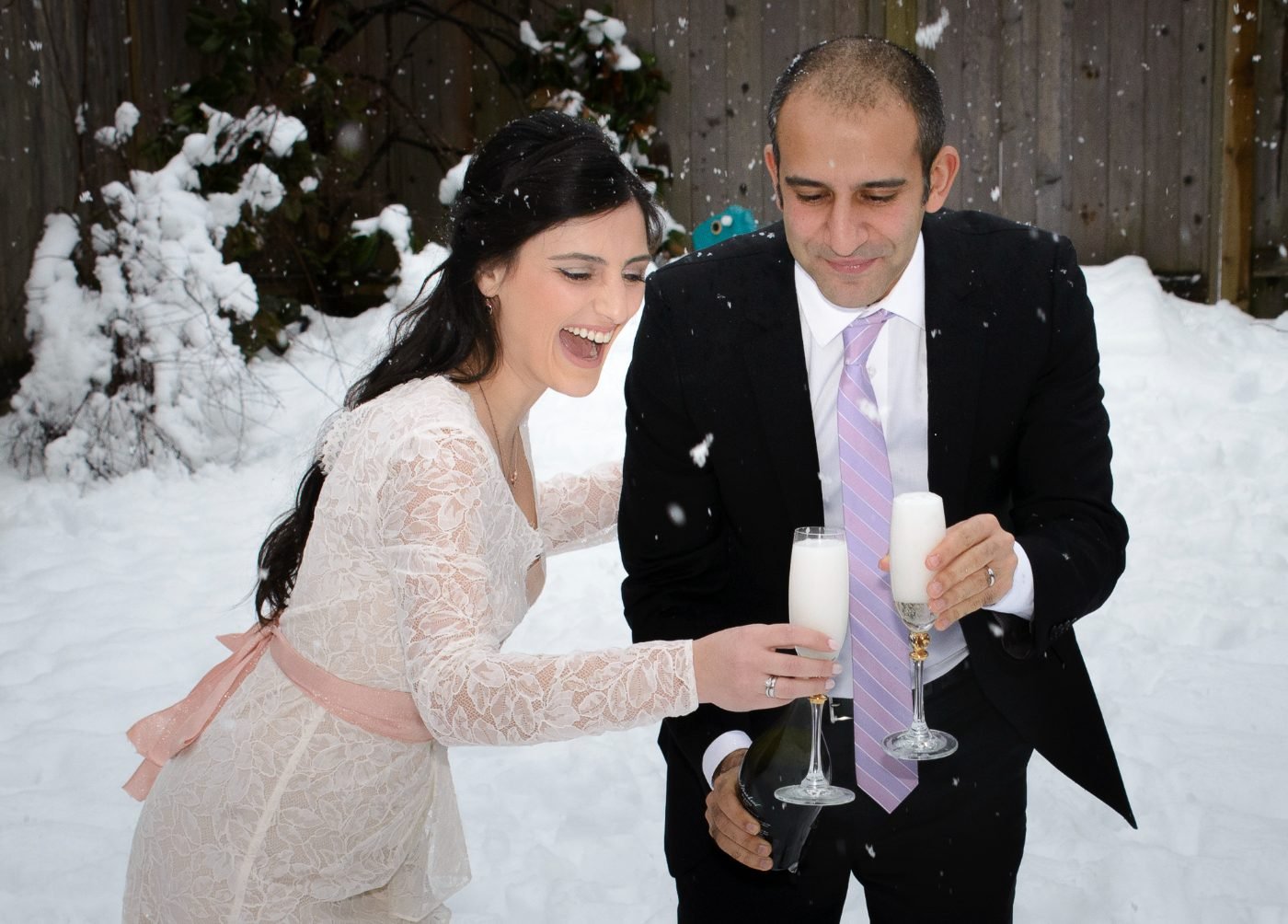 Bride and groom laughing as they toast with champagne in falling snow after their winter wedding