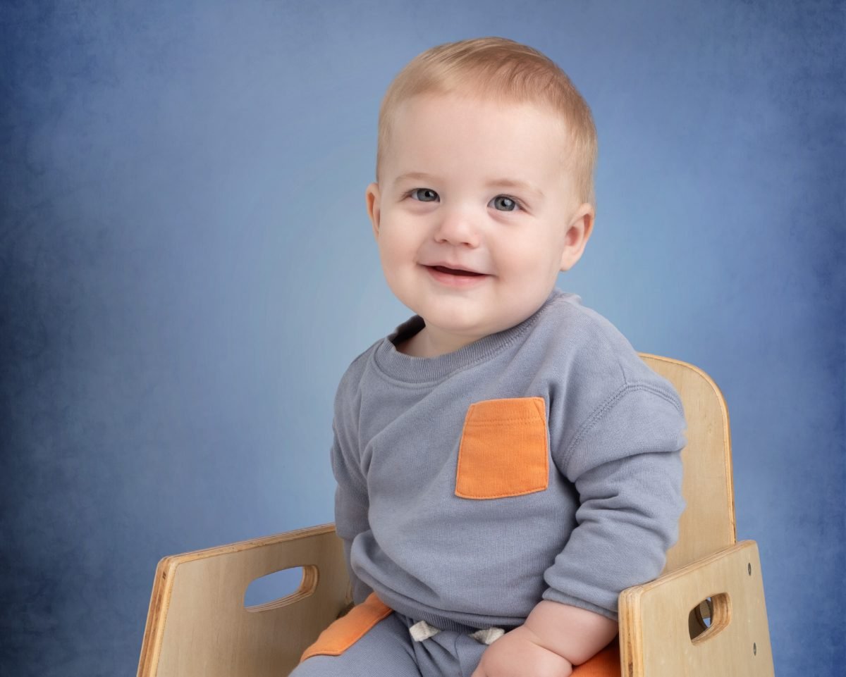 A toddler boy in a grey sweater sitting in a small wooden chair