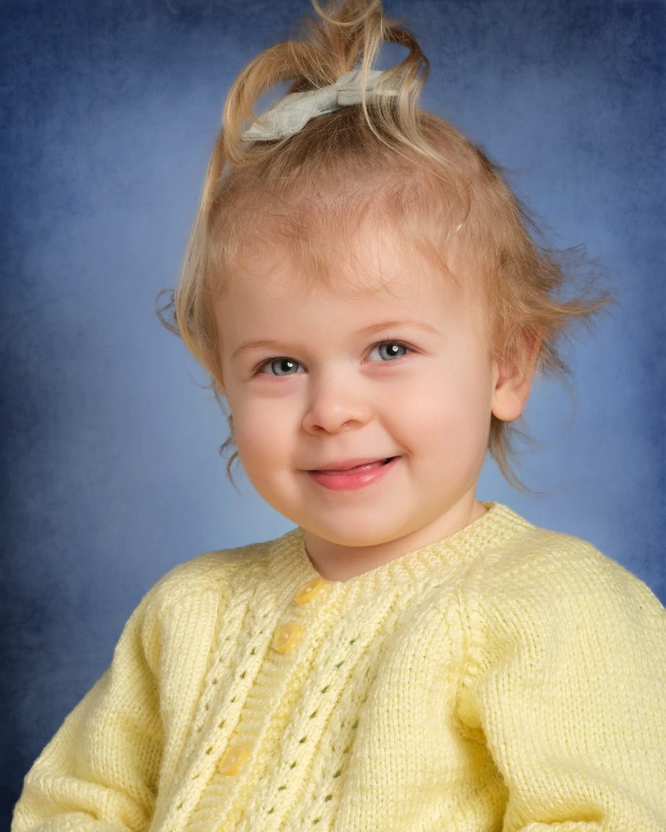 A toddler girl in a yellow cardigan on a classic mottled blue backdrop