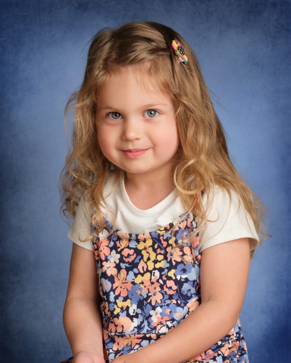 A student in a floral pinafore photographed on a classic mottled blue school-photo backdrop