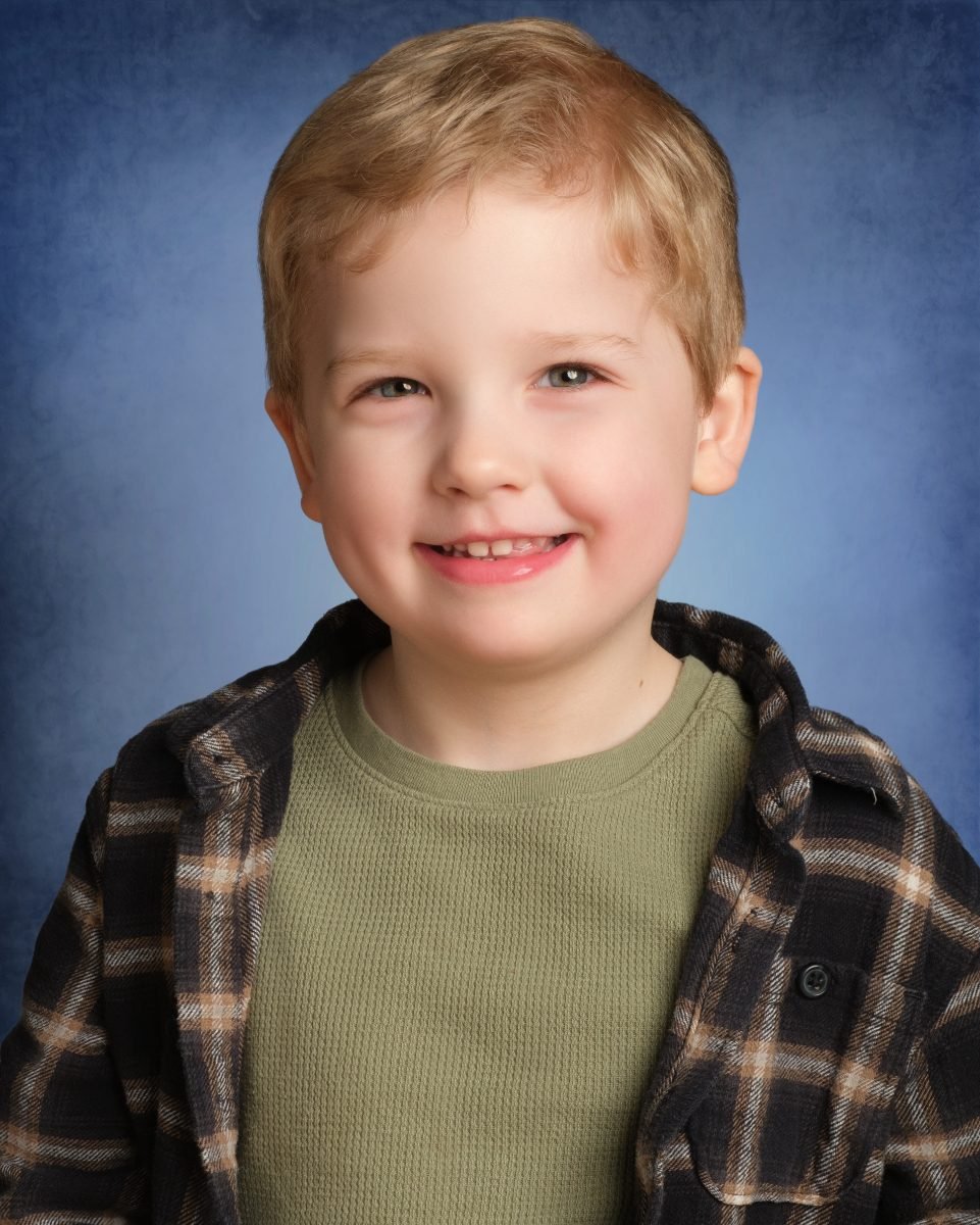 A preschool boy in a plaid shirt and olive tee smiling on a blue studio backdrop