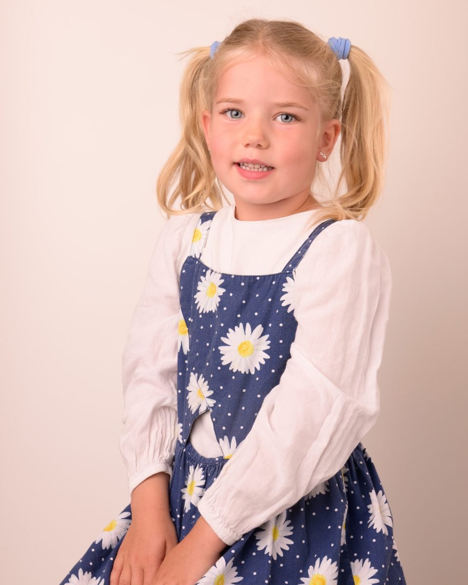 A girl with pigtails in a daisy pinafore on a warm neutral backdrop
