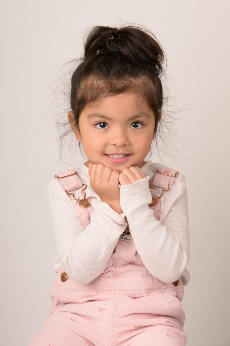 A girl with a top-knot resting her chin on her hands, warm neutral backdrop