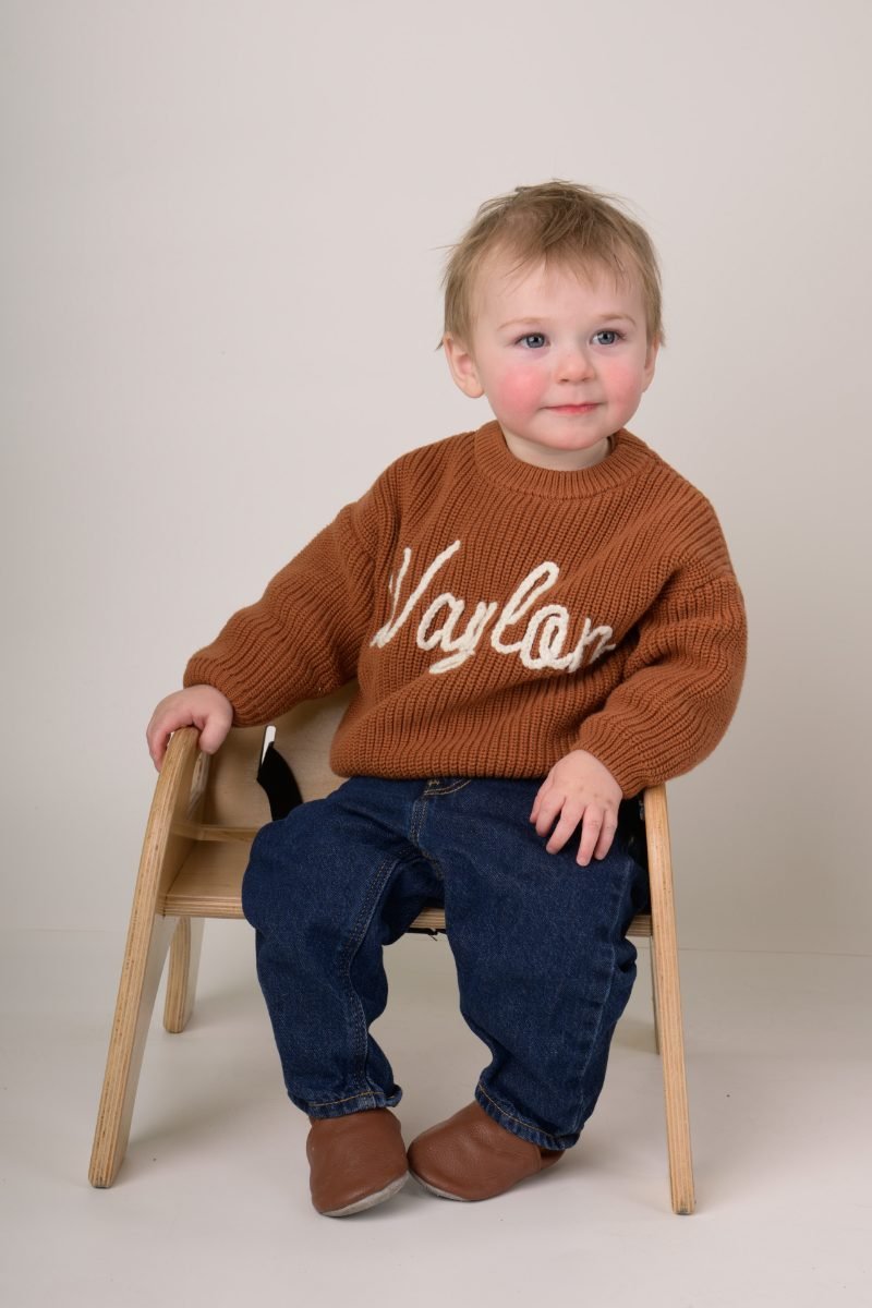 A toddler boy in a rust knit sweater sitting on a small wooden chair