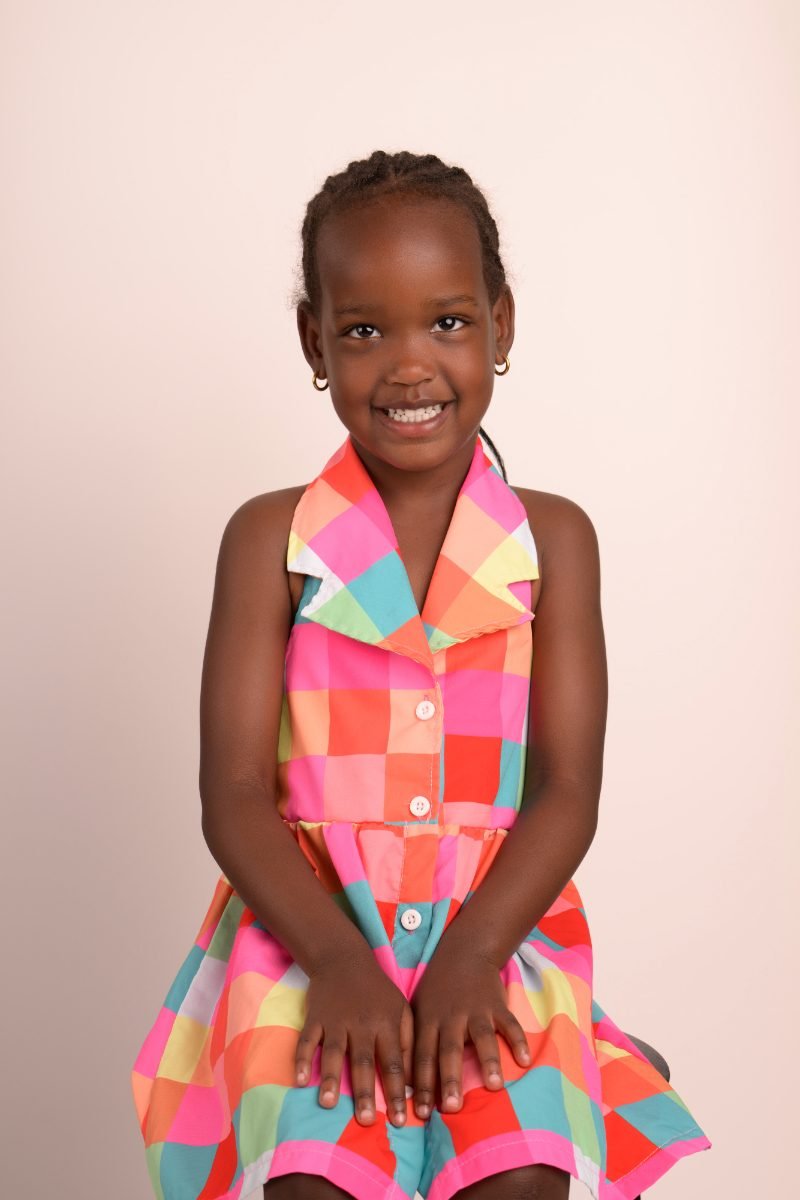A preschool girl in a rainbow check dress with her hands folded, smiling softly