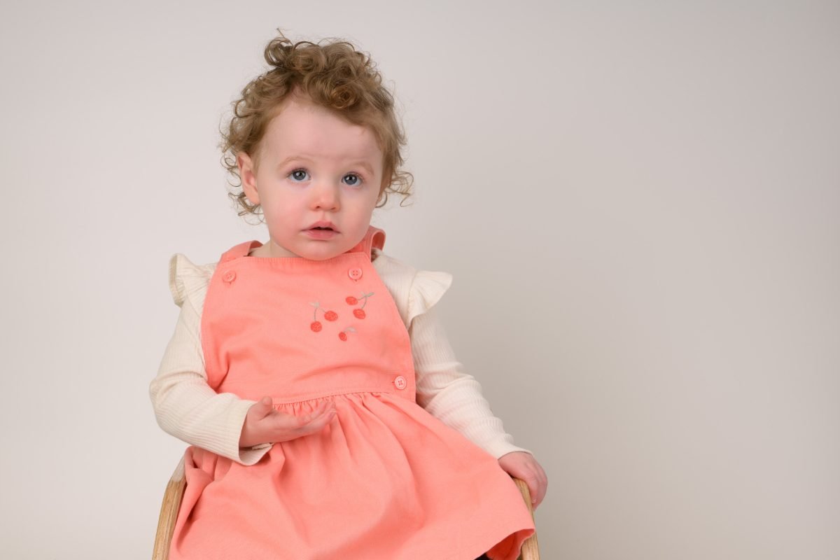 A curly-haired toddler in a coral dress sitting down