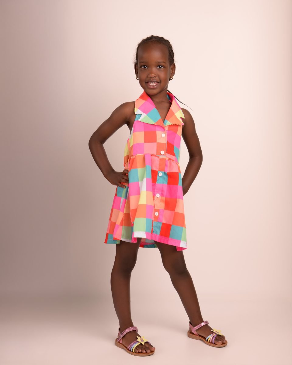 A girl in a rainbow check dress photographed head to toe, hand on hip