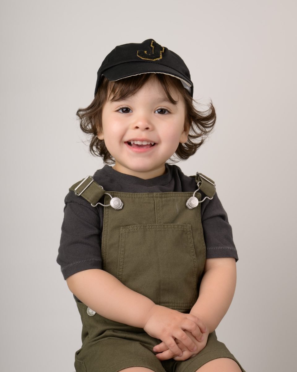 A toddler boy in a black cap and khaki overalls with his hands together