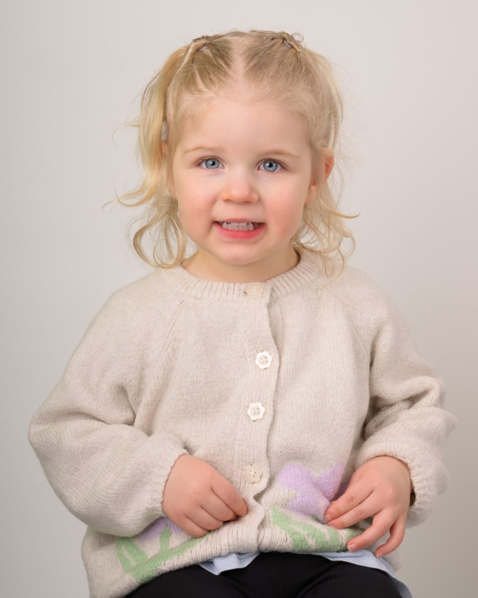 A toddler girl in a cream cardigan on a soft grey backdrop