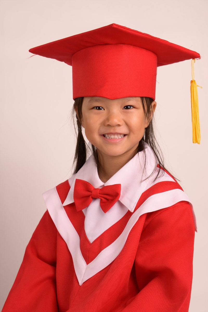 A preschool graduate in a red cap and gown with a bow-tie stole