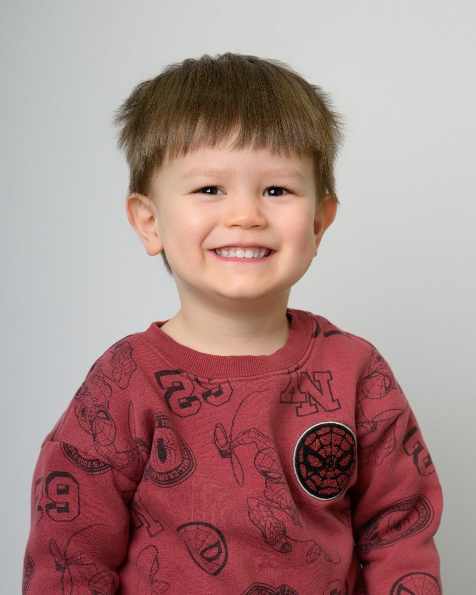 A toddler boy in a red patterned sweater grinning at the camera