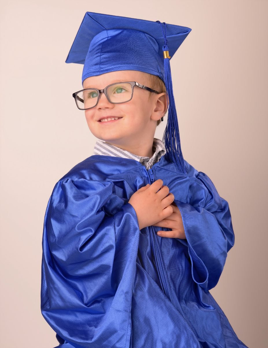 A preschool graduate in glasses and a royal blue gown, hand on his chest