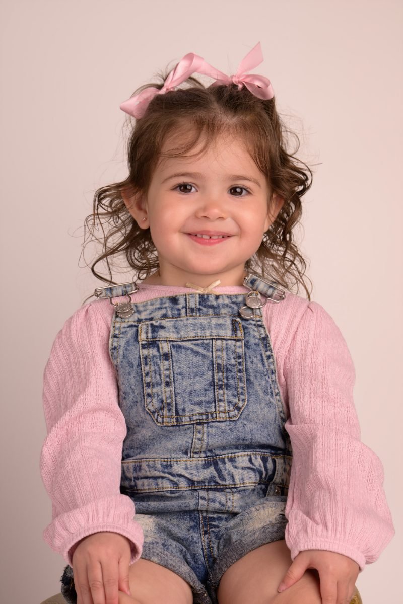 A girl in a denim pinafore and pink cardigan on a warm neutral backdrop