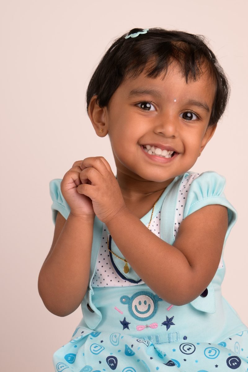 A girl in a teal dungaree top with her hands clasped at her shoulder