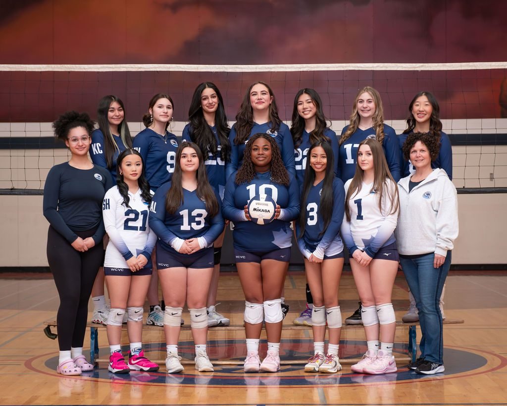 Team photo of a girls volleyball squad with their head coach in the gym